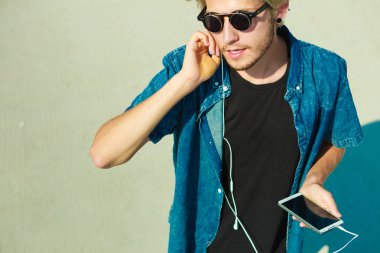 Tatil, dinlenme, tutku kavramı. Rahatlatıcı, smartphone cep telefonu ile müzik dinlemek ve güzel, güneşli havanın tadını güneş gözlüğü adam sarışın genç moda. Açık atış