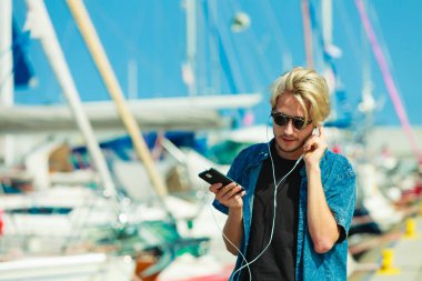 Tatil, rahatlama, tutku konsepti. Güneş gözlüklü genç, modaya uygun sarışın adam dinleniyor, müzik dinliyor ve güzel, güneşli havanın tadını çıkarıyor. Açık hava çekimi