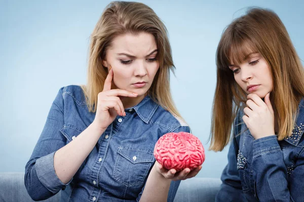 Friendship, human relations concept. Two women friends or sisters wearing jeans shirts, thinking about solving problem holding fake brain.