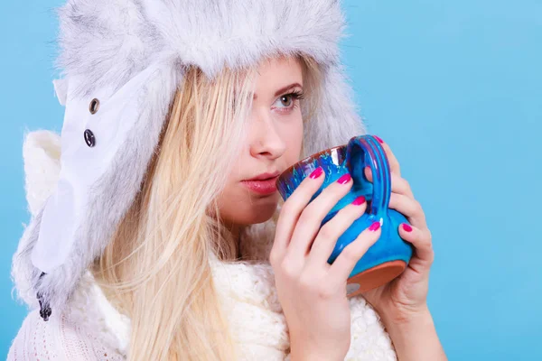 Accessories and clothes for cold days, fashion concept. Blonde woman in winter warm furry hat in russian style and scarf drinking hot drink from mug.