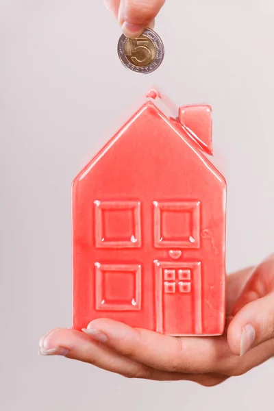 Household savings and finances, economy concept. Person putting money coin into moneybox in shape of house, studio shot on grey background