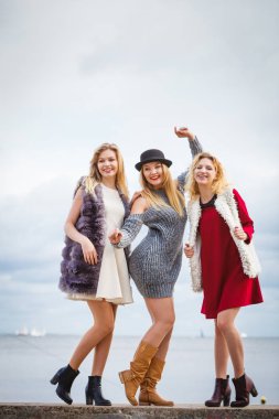 Üç modaya uygun kadın denizin kenarında şık kıyafetler giyiyorlar. Tarz, moda, arkadaşlık konsepti.