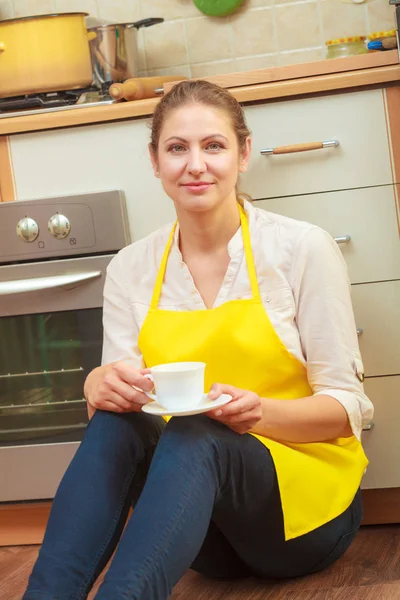 Mature woman in apron holding cup of coffee in kitchen. Housewife female relaxing resting sitting on floor.