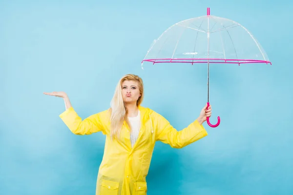 Blonde woman wearing yellow raincoat holding transparent umbrella checking weather if it is raining.