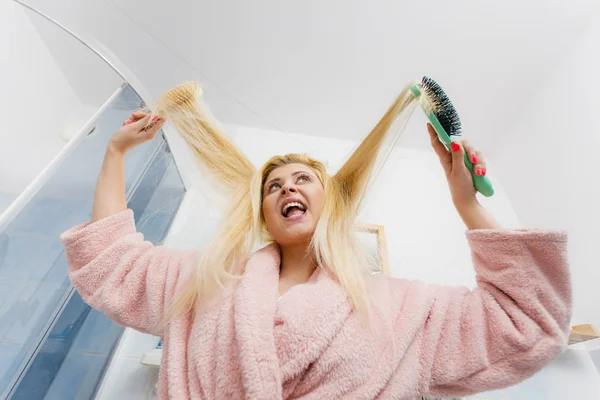 女人穿着睡袍试图刷她长长的金色很乱了头发 早上常规的美容保养 护发问题和发型设计概念 — 图库照片