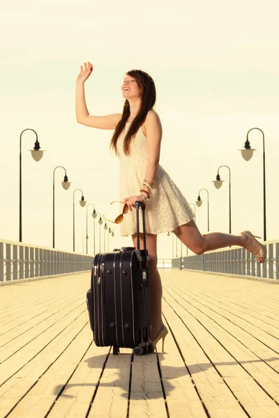 Travel, packing, journey concept. Woman wearing white short dress standing next to her suitcase waving to somebody, pier in background.