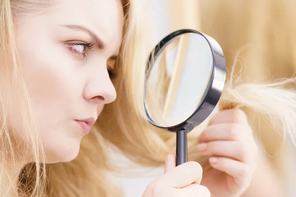 Split ends problem, dry effect, haircare concept. Unhappy blonde woman looking at destroyed damaged hair through magnifying glass
