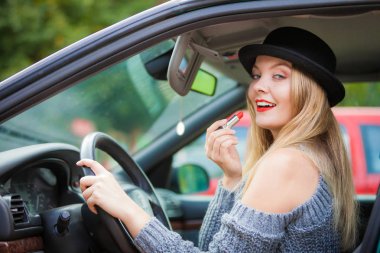 Dikiz aynasına bakan, dudaklarını boyayan, arabayı sürerken makyaj yapan çekici genç bir kadın..