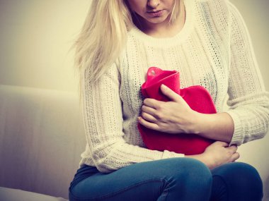 Acı dolu dönemler ve adet krampı sorunları konsepti. Karnı ağrıyan bir kadın kahve içerken kendini kötü hisseder sıcak su şişesini tutar ve biraz rahatlar.