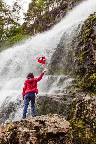 Tourist woman with norwegian flag at Svandalsfossen in Norway, powerful waterfall in norwegian mountains. National tourist Ryfylke route.