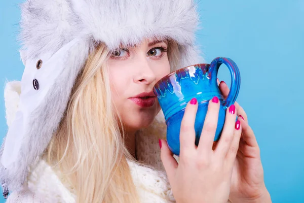 Accessories and clothes for cold days, fashion concept. Blonde woman in winter warm furry hat drinking hot drink from mug. Blue background.