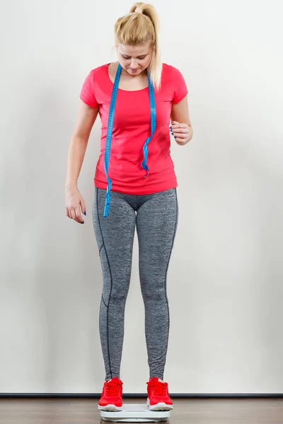 Healthy fit life style, controling body concept. Woman wearing sportswear, leggings and trainers standing on weight machine holding measuring tape.