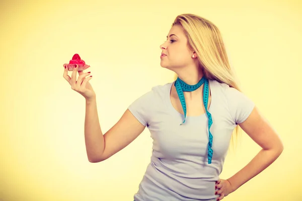 Restrictive diet, temptation, losing weight concept. Woman holding strawberry sweet cupcake having measure tape around her neck. Studio shot on yellow background