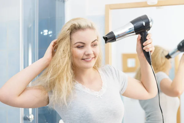 Positive woman using hair dryer on her blonde hairdo. Haircare, hairstyling concept.