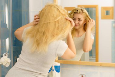 Yağlı saçlarıyla sorunu olan sarışın bir kadın banyoda kendine bakıyor. Kadın kafa derisini gösteriyor, kaşınıyor, kepek problemi var..