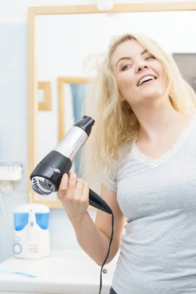 Positive woman using hair dryer on her blonde hairdo. Haircare, hairstyling concept.