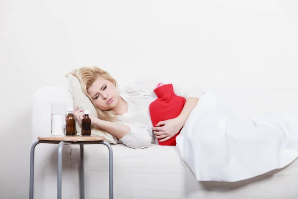 Painful periods and menstrual cramp problems concept. Woman having stomach cramps lying on cofa feeling very unwell holding hot water bottle to feel some relief