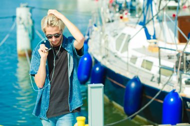 Tatil, rahatlama, tutku konsepti. Güneş gözlüklü genç, modaya uygun sarışın adam dinleniyor, müzik dinliyor ve güzel, güneşli havanın tadını çıkarıyor. Açık hava çekimi