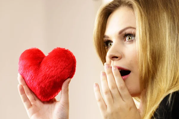 Funny woman realize falling in love. Young pretty woman having shocked face expression holding small heart shaped pillow.