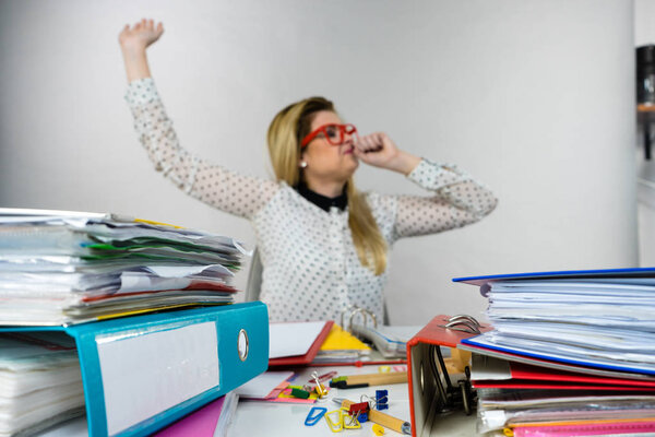 Sleepy business woman doing her work sitting working at desk full off documents in binders yawning being bored or tired staying up late.