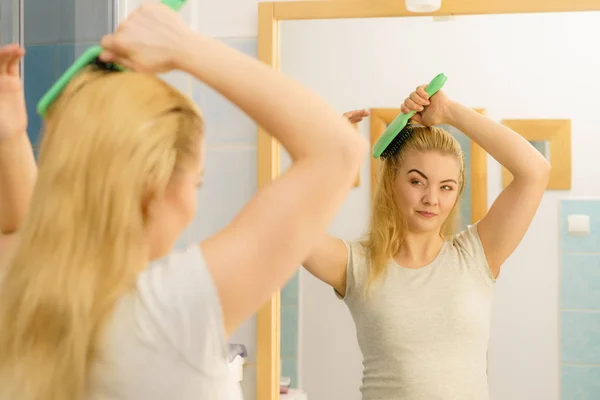 Happy woman combing her hair with brush. Young smiling female with natural blond straight long hairs in bathroom after morning shower.