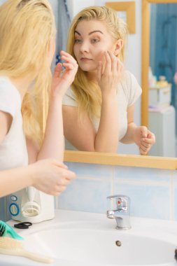 Banyo aynasında kendine bakan oldukça çekici genç bir kadın dış görünüşünü düşünüyor cilt durumunu kontrol ediyor.