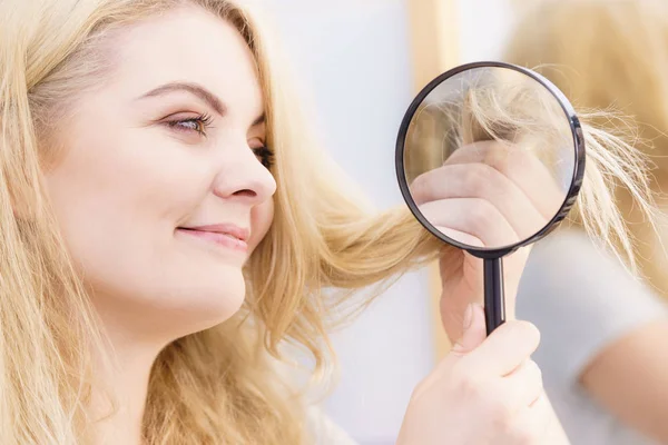 Happy woman magnifying her hair ends looking through magnifer being positive about conditioner results on her hairdor.