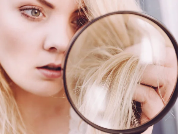 Haircare, bad effects of bleaching concept. Unhappy woman looking at ends of her blonde hair through magnifying glass