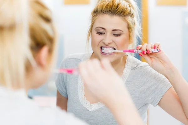 Woman brushing cleaning teeth closeup. Blonde girl with toothbrush in bathroom, looking at mirror. Oral hygiene.
