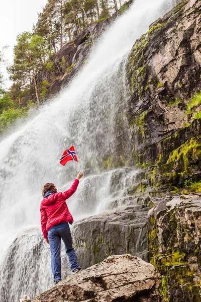 Tourist woman with norwegian flag at Svandalsfossen in Norway, powerful waterfall in norwegian mountains. National tourist Ryfylke route.