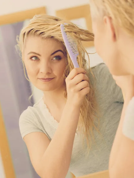 Pretty young woman taking care of haircare, brushing wet blonde hair after taking a shower feeling fresh.