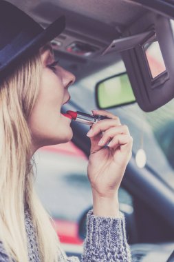 Dikiz aynasına bakan, dudaklarını boyayan, arabayı sürerken makyaj yapan çekici genç bir kadın..