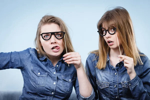 Elinde sahte gözlük olan iki mutlu kadın kot gömlek giyerek eğleniyor. Fotoğraf ve karnaval komik aksesuar konsepti.