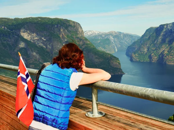 Female tourist with norwegian flag enjoying scenic view over fjord Aurlandsfjord from Stegastein viewpoint, take photos with camera. National tourist route Aurlandsfjellet.
