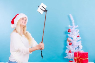 Noel hediyelerinin tadını çıkarıyorum. Noel Baba şapkalı bir kadın selfie çubuğu kullanarak kendi fotoğrafını çekiyor. Mavi arkaplanda kapalı çekim