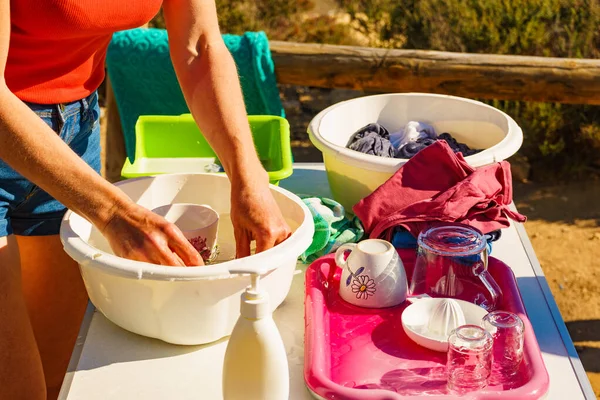 Mature woman washing up dishes in bowl on fresh air at camper car. Dishwashing outdoor on camping site, sea shore