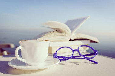 Tatillerde kitap okumak. Kahve fincanı ve kitaplar açık havada mavi deniz suyuna karşı. Kitap sayfaları rüzgarda çırpınıyor.