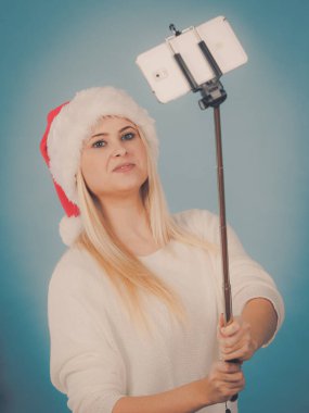 Noel hediyelerinin tadını çıkarıyorum. Noel Baba şapkalı bir kadın selfie çubuğu kullanarak kendi fotoğrafını çekiyor. Mavi arkaplanda kapalı çekim