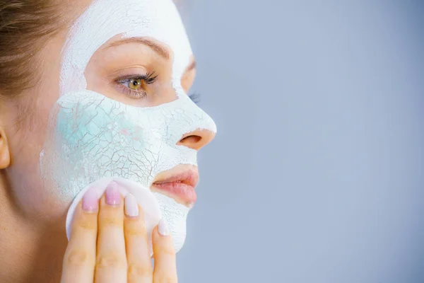 Young woman with white dried mud mask on face being removing cosmetic with cotton swab. Teen girl taking care of oily skin. Beauty treatment. Skincare.