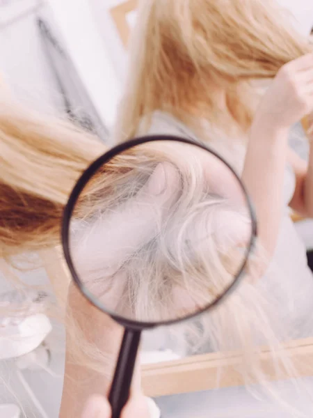 Haircare, bad effects of bleaching concept. Woman looking at ends of her blonde hair through magnifying glass