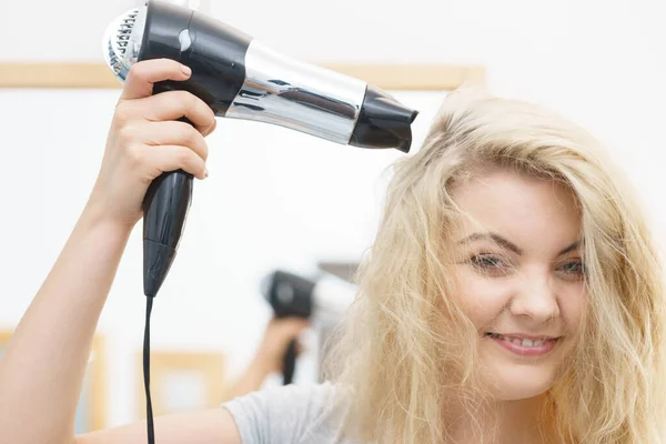 Positive woman using hair dryer on her blonde hairdo. Haircare, hairstyling concept.