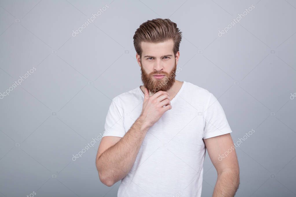 Studio shot of handsome pensive male holds chin, thinks about something, dressed in white t-shirt, stands against white background.Thoughtful bearded man