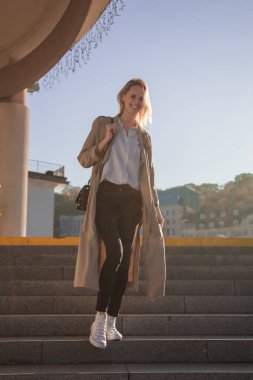 Genç kadın vintage binaların arka planı Caddesi boyunca yürümeye siyah deri çanta beyaz bir T-shirt moda bardaklarda şık bahar paltolu. Avrupa kız. Beyaz bir gömlek, siyah pantolon ve trençkot giymiş