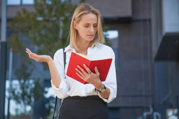 Happy blonde business woman with notebook against of office building. Dressed in a white shirt and black skirt