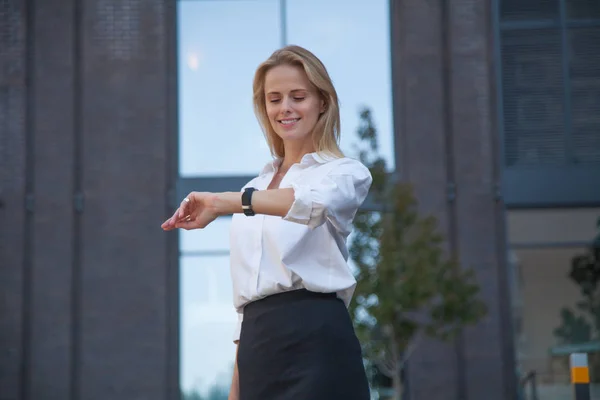 Happy blonde business woman checking time with watch on her hand against of office building. Dressed in a white shirt and black skirt