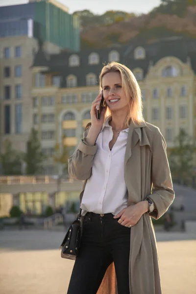 Una joven mujer de éxito en una gabardina, caminando por la calle, hablando por su teléfono celular . —  Fotos de Stock