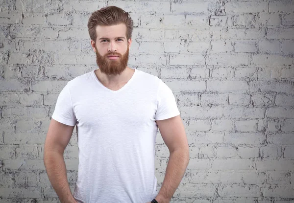 Serious thoughtful male with ginger beard, dressed casually, looking to the camera, isolated over white brick wall background.