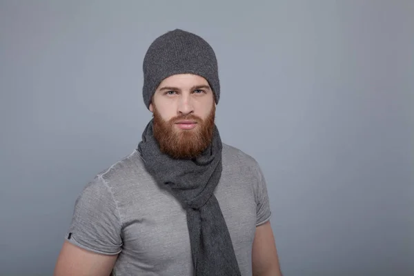 Joven hombre guapo con la barba en gorra gris y bufanda de pie junto a la pared gris y mirando directamente a la cámara —  Fotos de Stock