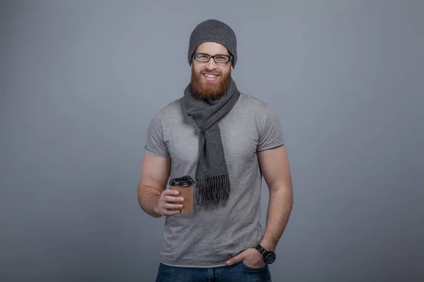 Jovem bonito homem com a barba em óculos vestindo chapéu cinza, cachecol e um chapéu em pé junto à parede de tijolo com uma xícara de café na mão e segurando outra mão no bolso — Fotografia de Stock