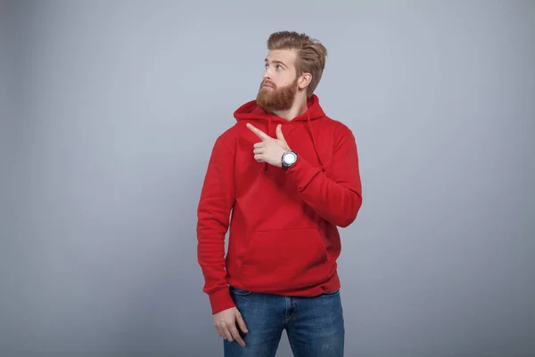 Joven hombre barbudo guapo vistiendo sudadera roja de pie sobre el fondo gris y señalando su dedo en el lado izquierdo y mirando allí con el interés —  Fotos de Stock